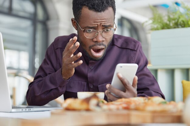 Frustrated dark skinned male looks desperately into screen, reads information on smart phone, sits in coffee shop