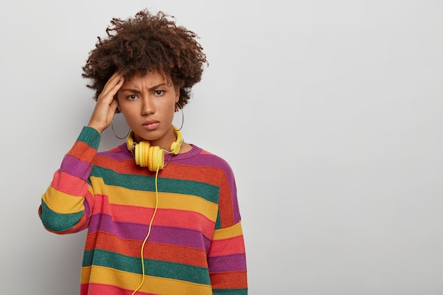 Frustrated dark skinned woman has headache after long listening of music, displeased face expression