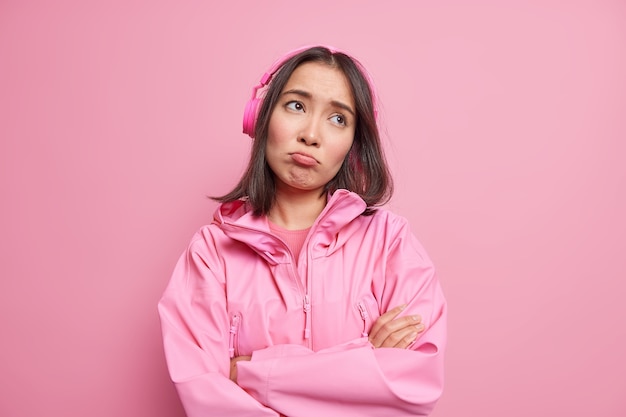 좌절 된 실망한 아시아 여성은 우울한 슬픈 표정을 가지고 팔을 접은 채 신중하게 집중하여 무선 헤드폰을 통해 음악을 듣고 분홍색 벽에 고립 된 재킷을 입습니다.