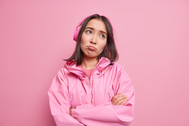 欲求不満の失望したアジアの女性は悲観的な悲しげな表情で腕を組んで思慮深く集中し続けますワイヤレスヘッドフォンで音楽を聴きますピンクの壁に隔離されたジャケットを着ています