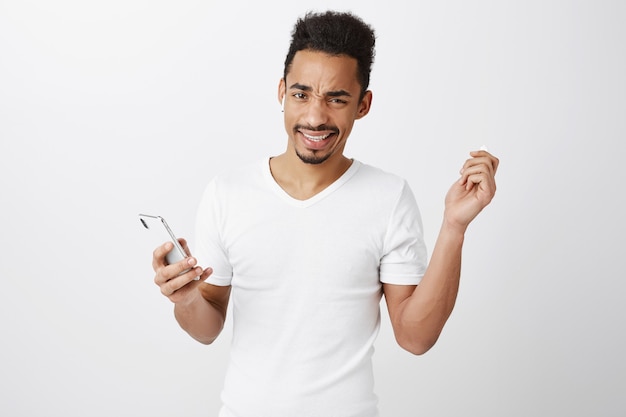 Frustrated and displeased african-american guy take-off earphone and cringe, holding mobile phone