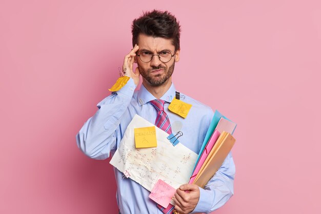 Frustrated displeased office worker has headache keeps hand on temple looks discontent at camera being tired because of paper work poses 