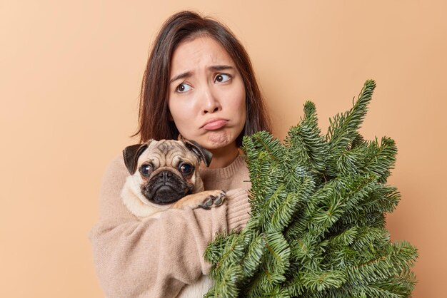 Frustrated displeased woman poses with pug dog and fir tree branches celebrates New Year alone feels lonely wears jumper poses against beige background. People winter holidays animals concept