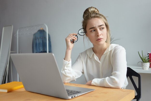 Frustrated exhausted young woman blogger dressed in white blouse holding round eyeglasses and looking away with worried expression, searching for inspiration, trying to write article for her blog