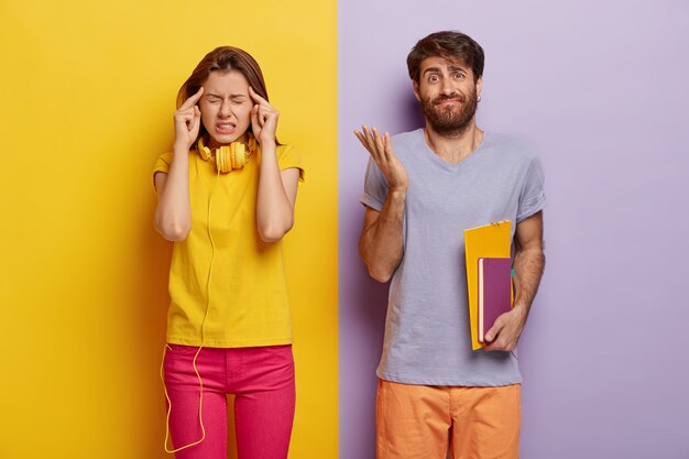 Frustrated female student keeps index fingers on temples, suffers from headache, doubtful male groupmate holds notebooks