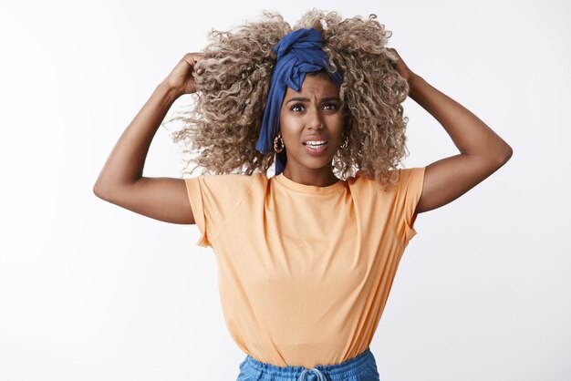 Frustrated and furious emotive africanamerican blond woman afro hairstyle stylish outfit cringe from shock and disappointment hold hands on hair pissed staring troubled and questioned camera