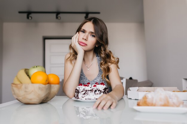 ケーキと果物を持ってテーブルに座っている退屈な若い女性 朝食時にポーズをとるかわいい白人の女の子 無料の写真