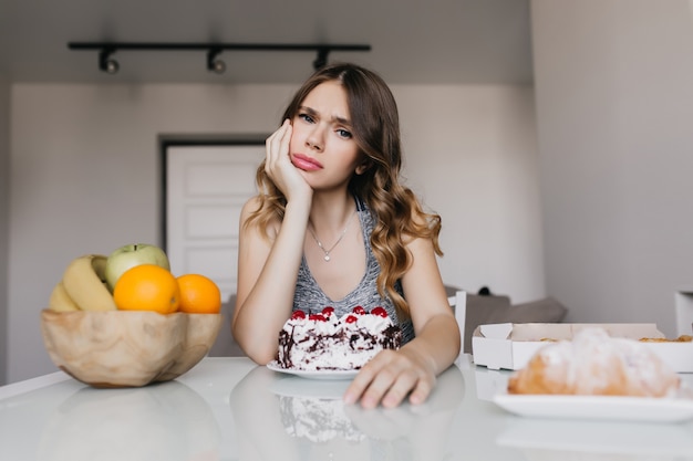 Frustrated girl posing with cake in morning. Indoor shot of european woman eating fruits during diet.