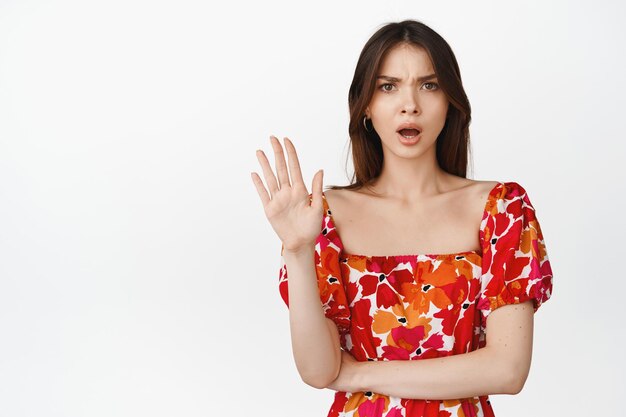 Разочарованная девушка показывает стоп-жест, машет рукой, чтобы отклонить что-то, говорит, что не стоит злиться на белом фоне в красном цветочном платье