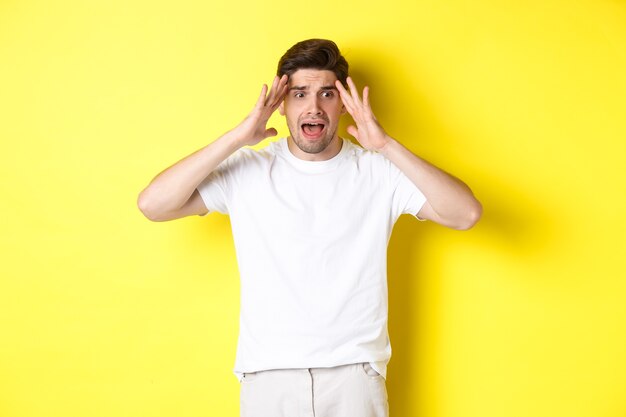 Frustrated guy looking shocked, panicking and holding hands on head, standing against yellow