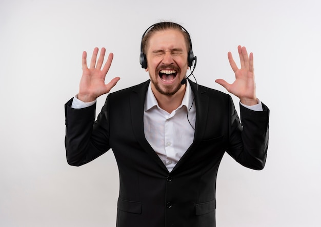 Frustrated handsome businessman in suit and headphones with a microphone shouting and yelling with aggressive expression standing over white background