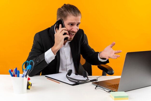 Разочарованный красивый бизнесмен в костюме, работающий на ноутбуке, разговаривает по мобильному телефону, выглядит смущенным и недовольным, сидя за столом в офисе на оранжевом фоне