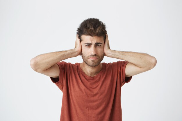 Frustrated handsome hispanic man in red tshirt plugging ears with hands being exhausted of loud sounds from neighboring apartments at night.