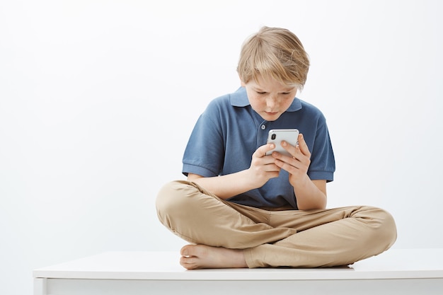 Free photo frustrated intense cute young boy with blond hair in casual outfit, sitting on crossed feet