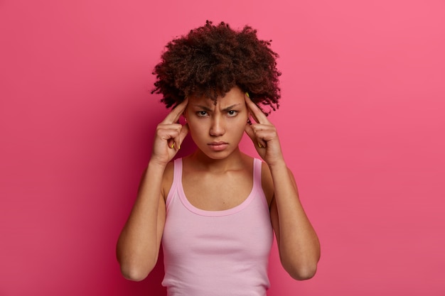 Frustrated intense ethnic woman touches temples, tries to focus on something, cannot concentrate because of headache, looks seriosly , wears casual shirt, isolated on pink wall