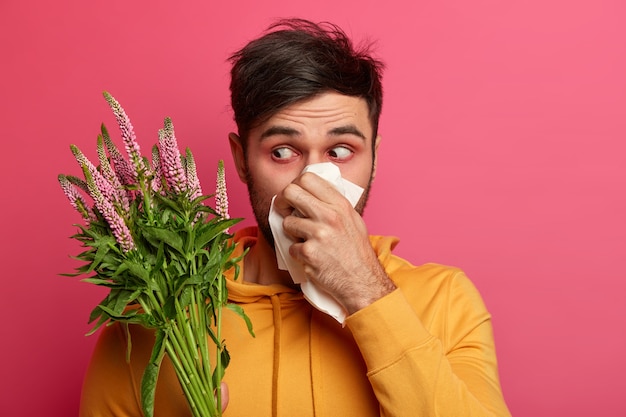 L'uomo frustrato soffia il naso nei tessuti, ha arrossamenti intorno agli occhi, sintomi di allergia, ha un aspetto malsano, concentrato sul fiore che sboccia, soffre di rinite, reazione allergica. persone e malattie