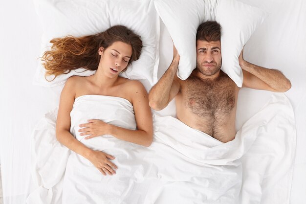欲求不満の男は枕で耳を覆い、大声で妻がいびきをかくために眠ることができず、不眠症に苦しみ、寝室でポーズをとる。いびきをかく女性にイライラする疲れた男。人、健康、睡眠障害