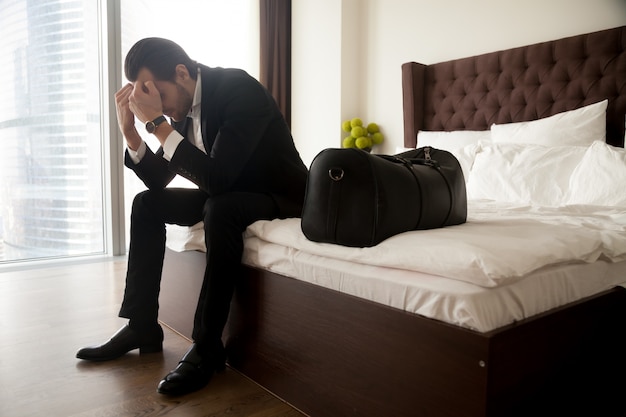 Frustrated man in suit sitting on bed besides luggage bag.