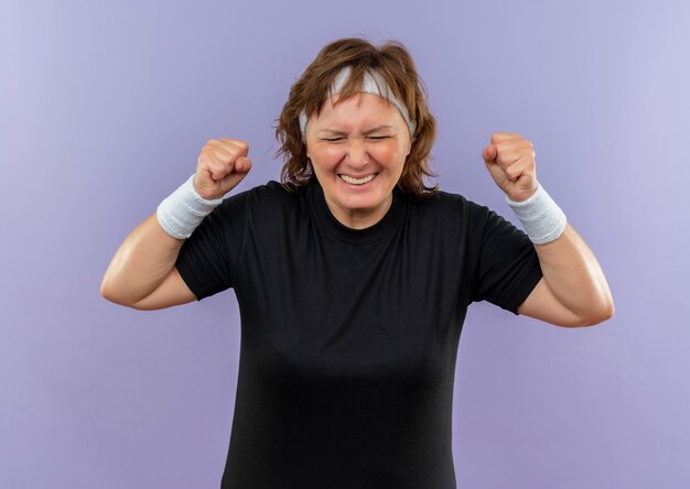 Бесплатное фото Разочарованная спортивная женщина средних лет в черной футболке с повязкой на голове, сжимая кулаки с раздраженным выражением лица, сумасшедшая, безумная, стоит у синей стены