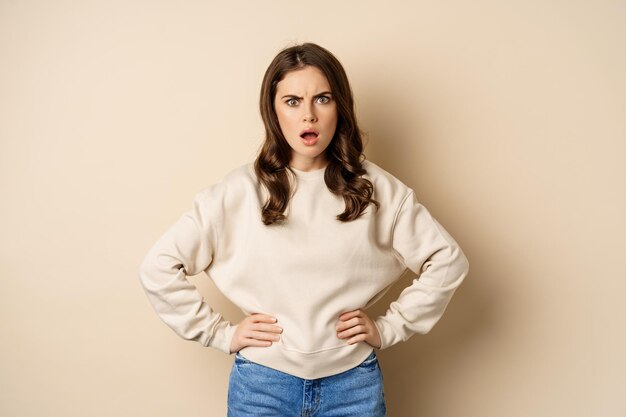 Frustrated, offended young woman looking hurt ad insulted at camera, standing shocked over beige background