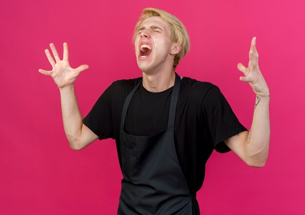 Frustrated professional barber man in apron shouting with aggressive expression with raised arms crazy mad 