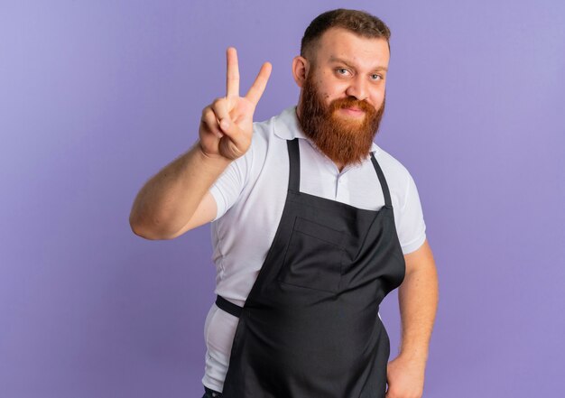 Frustrated professional bearded barber man in apron smiling showing victory sign standing over purple wall