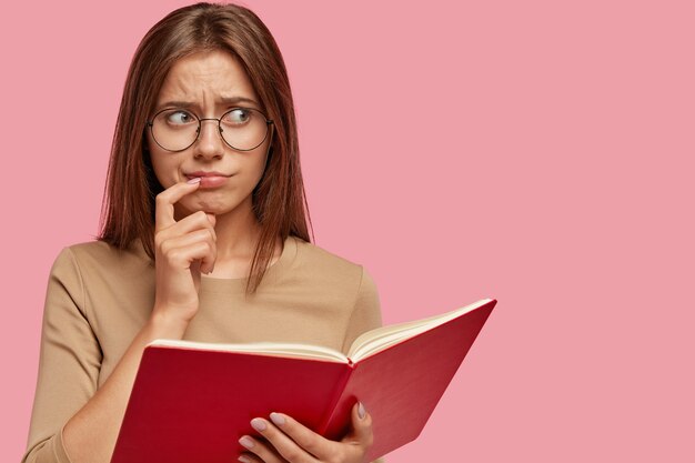 Frustrated puzzled schoolgirl keeps index finger near lips, carries red book, thinks in bewilderment