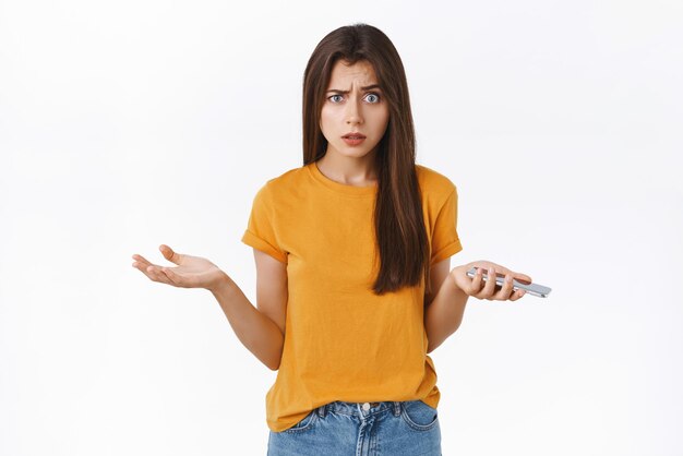 Frustrated puzzled seriouslooking shocked woman in yellow tshirt hold smartphone in spread hands and staring camera gasping stunned receive strange message standing white background