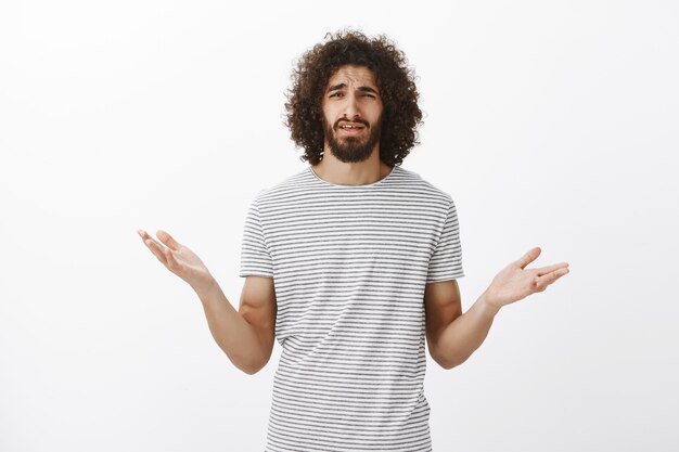 Frustrated questioned handsome man with beard and curly hair, spread hands cluelessly