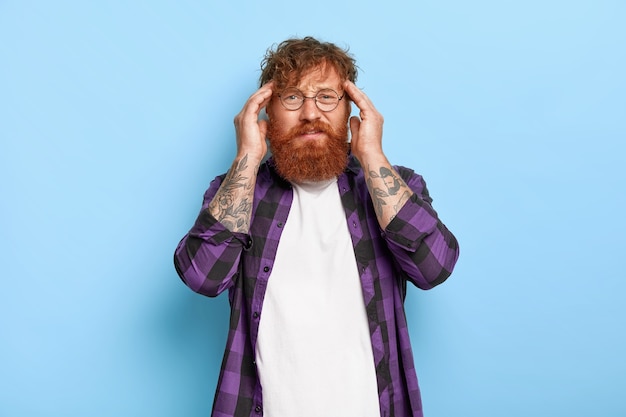 Foto gratuita un uomo frustrato dai capelli rossi con una folta barba tocca le tempie, soffre di un'enorme emicrania, ha bisogno di antidolorifici