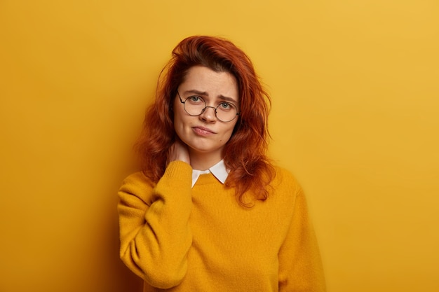 Frustrated red haired woman has strong pain in neck after working long time at computer, looks sadly at camera, suffers from osteohondrosis, has gloomy expression