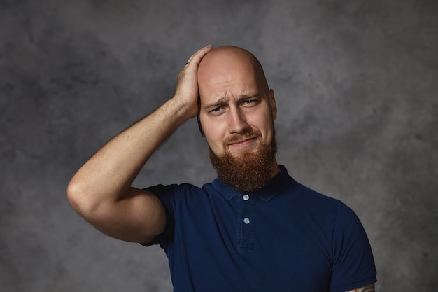 Frustrated regretful stylish young male with thick beard having forgetful puzzled expression, touching his shaved head, trying to remember something. Bearded guy suffering from terrible headache