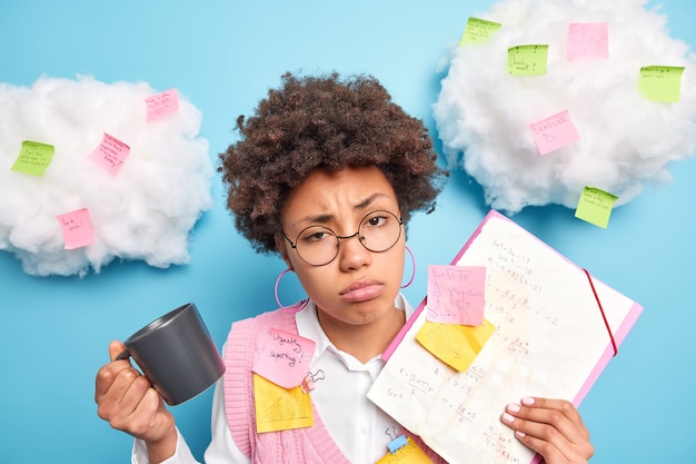 Free photo frustrated sad afro american student has tired expression feels sleepy after praparing for exam all day long surrounded by pasted stickers makes notes on papers to remember information drinks coffee