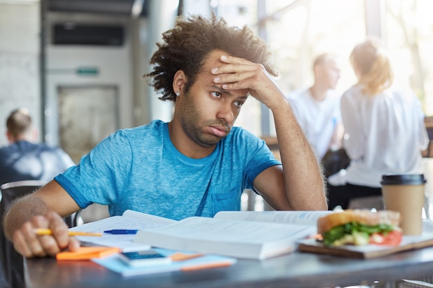 Studente maschio stressato frustrato seduto al tavolino del bar con libri, appunti e pranzo, con uno sguardo stanco esausto mentre non riesce a risolvere il problema matematico.