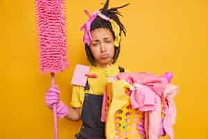 Free photo frustrated tired housewife holds mop and basket of laundry feels exhausted of doing cleaning at home