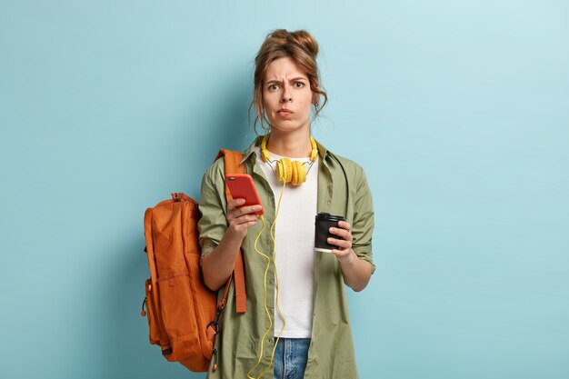 Разочарованная уставшая девушка из поколения миллениума пьет кофе на вынос, держит смартфон, подключенный к наушникам, наслаждается плейлистом