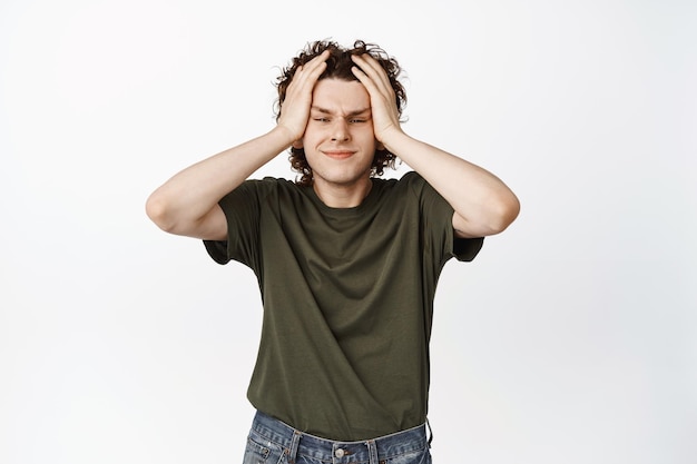 Frustrated and troubled young teen guy holding hands on head staring distressed and upset at camera standing in green tshirt over white background