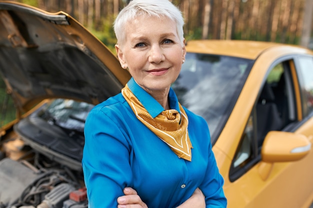 Pensionata femmina infelice frustrata in piedi vicino alla sua auto con il cofano aperto, con le braccia incrociate, in attesa di assistenza stradale.