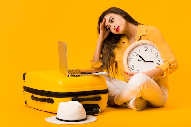 Frustrated woman holding clock and looking at laptop on top of luggage