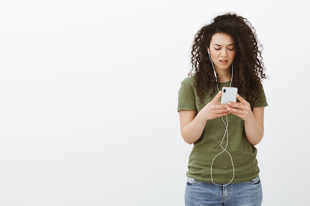 Frustrated woman staring at smartphone cluelessly. Portrait of confused displeased curly-haired woman in casual outfit