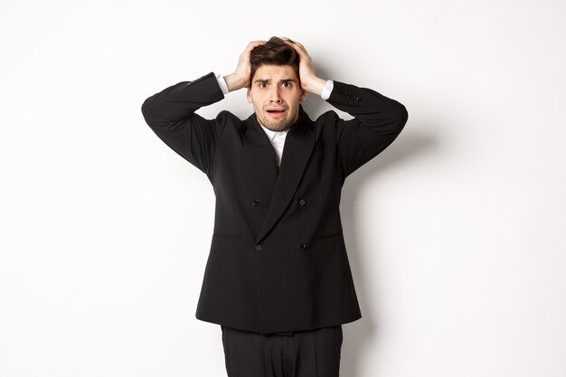 Frustrated and worried businessman in black suit, panicking as looking at trouble, holding hands on head alarmed, standing against white background