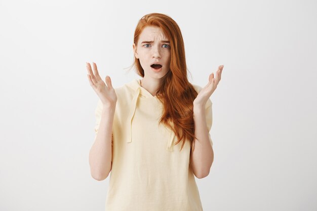 Frustrated worried redhead girl raise hands up in dismay, looking upset