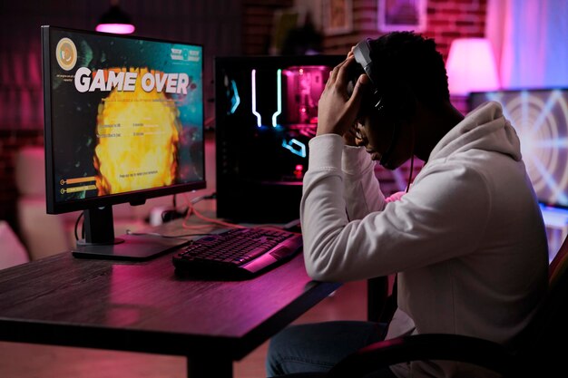 Frustrated young adult losing online video games play competition on computer with neon lights. Male gamer streaming action gameplay and feeling sad about lost shooting championship.