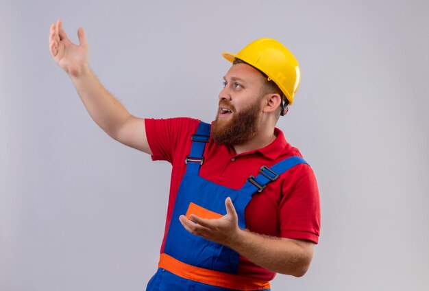 Frustrated young bearded builder man in construction uniform and safety helmet looking up with arms raised
