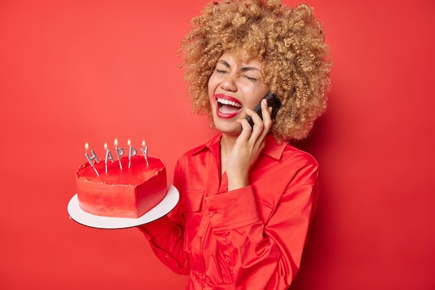 Разочарованная молодая кудрявая женщина разговаривает по телефону, плачет от отчаяния, носит рубашку, держит сердечный торт с горящими свечами, расстроенная, чтобы отпраздновать день рождения в одиночестве на красном фоне
