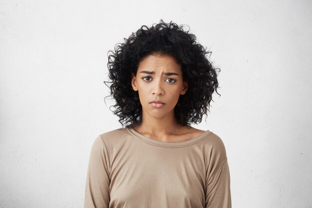 Frustrated young dark-skinned woman with shaggy black hair frowning, looking with sad and unhappy expression while going through hard times in her life. Human emotions and feelings