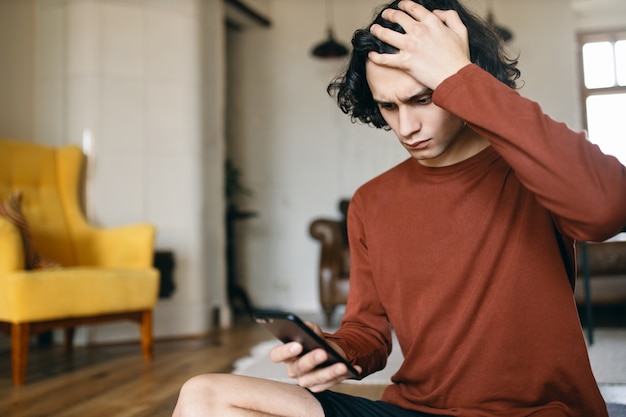 Frustrated young man having stressed facial expression, keeping hand on his head, holding cell phone, reading bad news