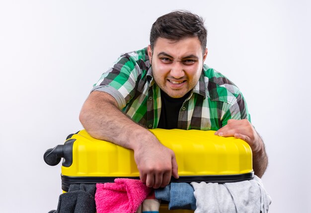 Frustrated young traveler man in checked shirt standing with suitcase full of clothes tryinbg to close it with angry face over white wall