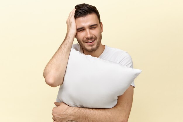 Frustrated young unshaven male embracing white pillow and holding hand on his head having hangover, suffering from headache because of sleepless night, having sleepy tired facial expression