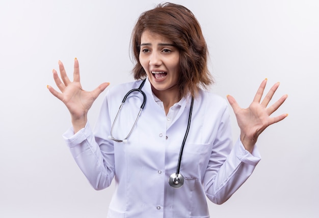 Frustrated young woman doctor in white coat with stethoscope rasing hands yelling with annoyed expression 